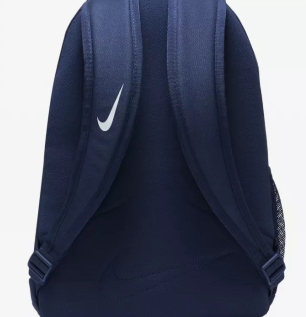 The back view of a navy blue Nike Rucksack backpack, showing padded shoulder straps, a white Nike swoosh logo on the left strap, and a mesh side pocket on a light gray background.